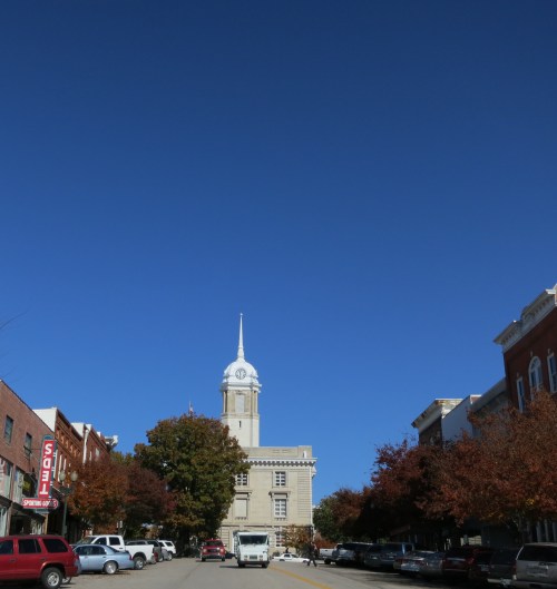 A street view featuring a clock tower, trees with autumn leaves, and clear blue skies.
