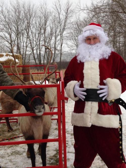 Santa Claus in a red suit stands next to a reindeer in a snowy setting, with trees in the background.