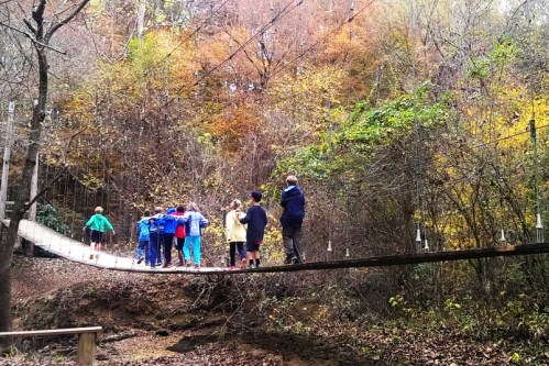 A group of children crossing a wooden bridge surrounded by autumn foliage in a forested area.