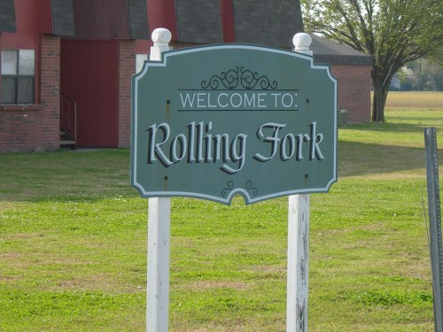 Welcome sign for Rolling Fork, set against a grassy landscape and a building in the background.