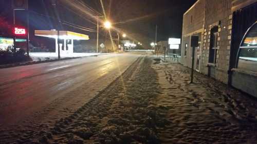 A snowy street at night, with a gas station and buildings illuminated by streetlights.