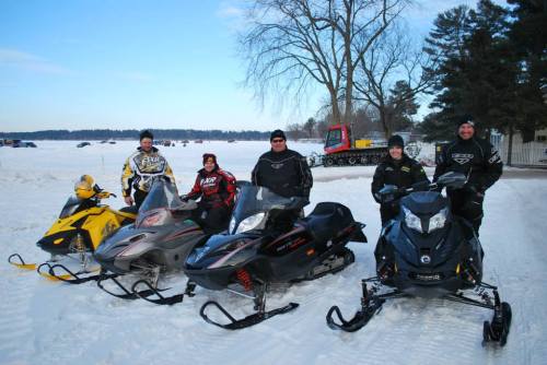 A group of six people stands beside snowmobiles on a snowy landscape, with trees and a frozen lake in the background.