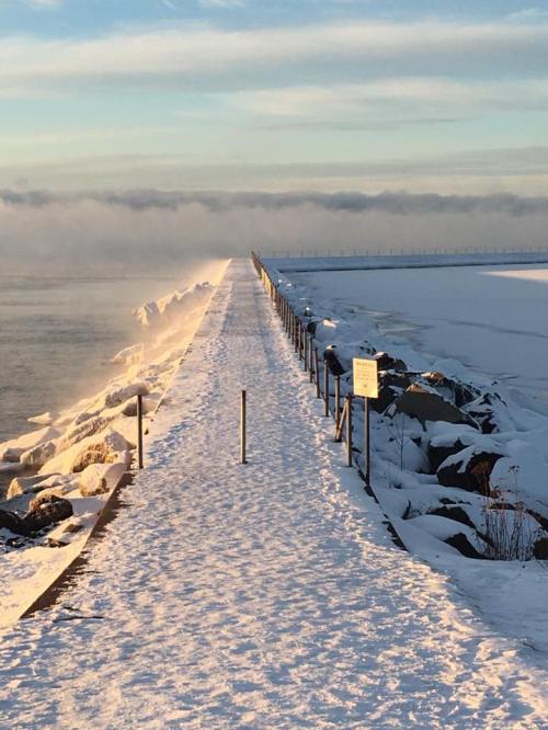 A snowy pathway leads to a foggy horizon, bordered by rocks and a fence, under a clear blue sky.