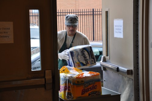 A person in military attire carries a box of napkins and toilet paper through a doorway.