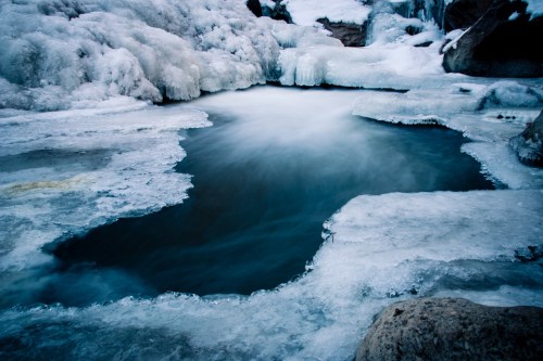 A serene, icy landscape featuring a partially frozen pool surrounded by snow-covered rocks and ice formations.