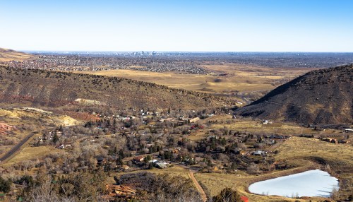 A panoramic view of a valley with a small town, fields, and a distant city skyline under a clear blue sky.