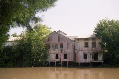 Abandoned, weathered building by a river, surrounded by lush greenery and reflecting in the water.