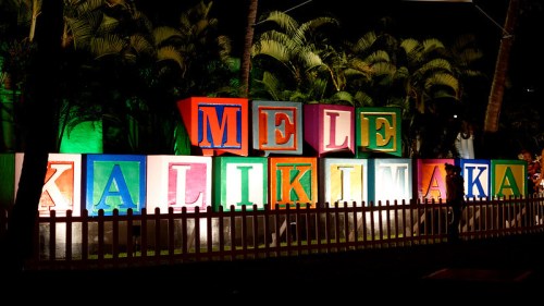 Colorful blocks spell "Mele Kalikimaka" against a nighttime backdrop with palm trees.