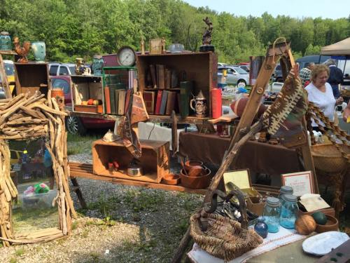 A vibrant outdoor market display featuring vintage items, books, and crafts, surrounded by greenery and shoppers.