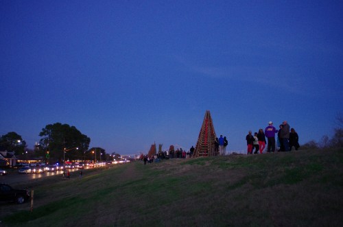 A crowd gathers on a grassy hill at dusk, with a large wooden structure and traffic visible in the background.