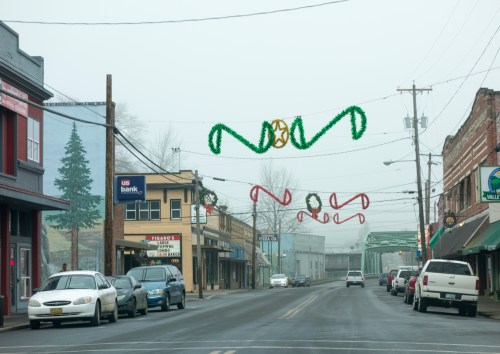 A foggy street scene decorated with holiday garlands, featuring shops and parked cars along the road.