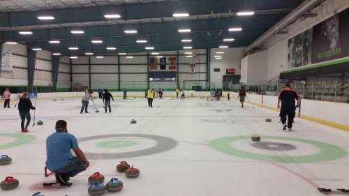 A curling rink with multiple players sliding stones on the ice, practicing and competing in a lively atmosphere.