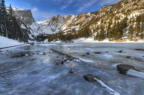 A serene winter landscape featuring a frozen lake surrounded by snow-covered mountains and evergreen trees.