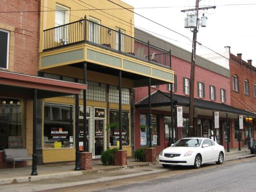 A street view of a small town with brick buildings, a balcony, and a parked white car.