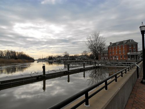 A calm river scene with a dock, birds resting, and buildings along the shore under a cloudy sky.