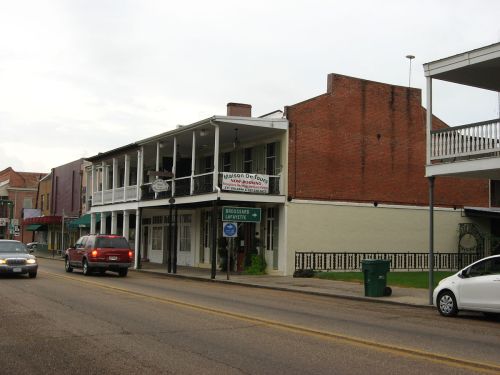 Historic buildings line a street, featuring balconies and storefronts, with cars parked along the road.