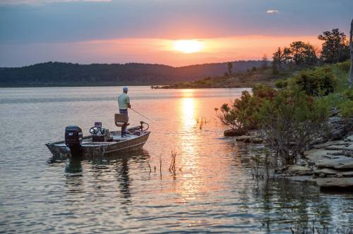 A person fishing from a small boat at sunset, with calm waters and a scenic landscape in the background.