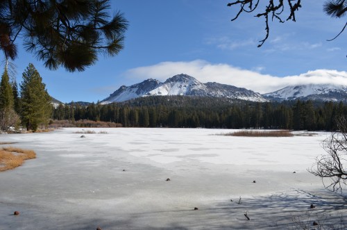 A serene winter landscape featuring a frozen lake, snow-capped mountains, and pine trees under a clear blue sky.