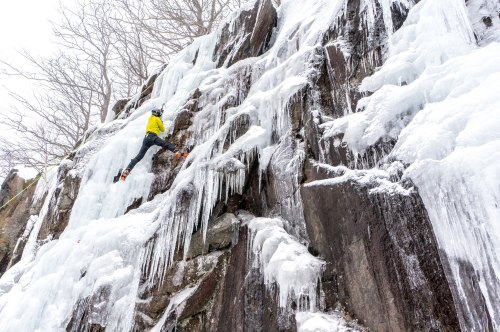 A climber ascends a frozen waterfall, using ice axes and crampons on a rocky, icy surface surrounded by bare trees.