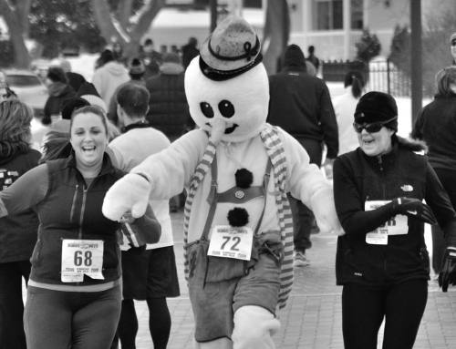 Two women smile while running alongside a snowman mascot in a lively outdoor event.
