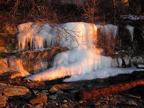 Icicles and frozen water cascade over rocky terrain, illuminated by warm sunset light.