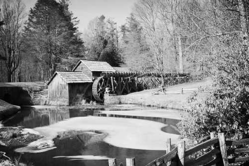 A historic watermill beside a pond, surrounded by trees, captured in black and white.