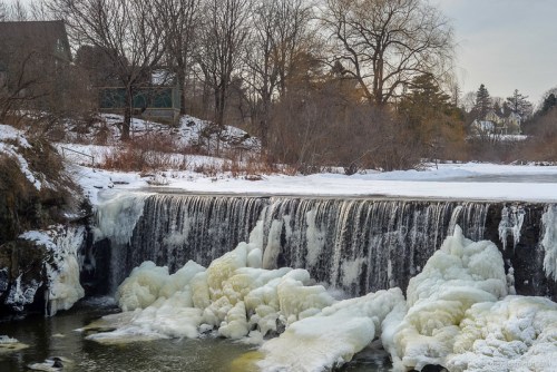 A frozen waterfall cascades over rocks, surrounded by snow-covered trees and a serene winter landscape.