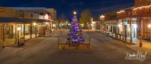 A festive street scene at night featuring a brightly lit Christmas tree and decorated storefronts.