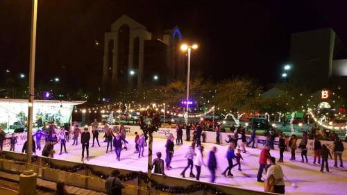 A lively outdoor ice skating rink at night, filled with skaters and festive lights in the background.