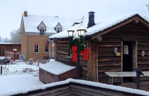 A snowy scene featuring a rustic log cabin with festive decorations and a nearby historic building in the background.