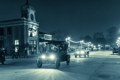 Two vintage cars drive through a dimly lit street, adorned with festive lights, in a nostalgic nighttime scene.