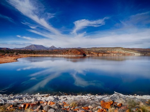 A serene lake reflects the sky and mountains, surrounded by rocky shores and vibrant vegetation.