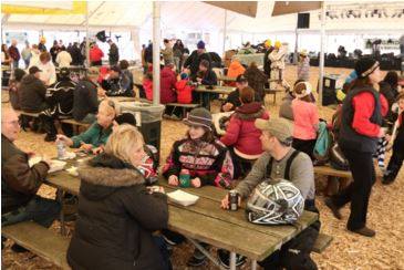 A busy indoor event with people sitting at tables, eating and socializing in a large tent.