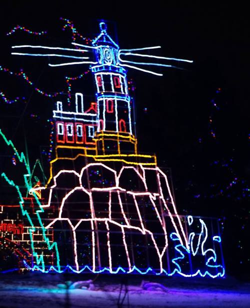 Colorful holiday lights depict a lighthouse on a rocky shore, surrounded by festive decorations and a dark night sky.