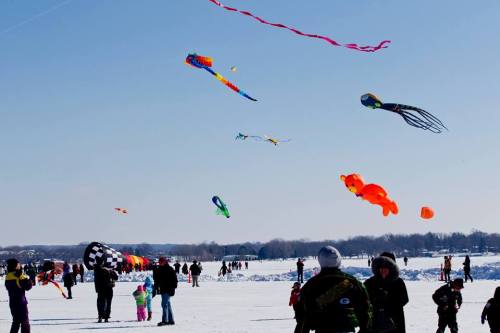 Colorful kites flying over a snowy landscape, with people enjoying a winter kite festival.