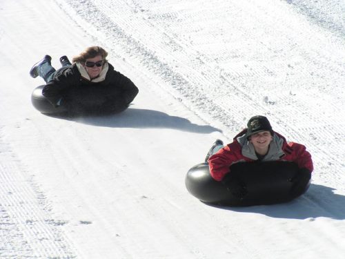 Two people sliding down a snowy hill on inner tubes, smiling and enjoying the winter fun.