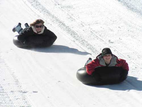 Two people sliding down a snowy hill on inner tubes, smiling and enjoying the winter fun.