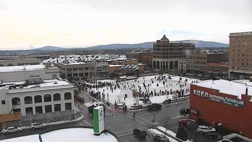 A snowy outdoor ice skating rink surrounded by buildings and mountains in the background. People are skating and enjoying the scene.