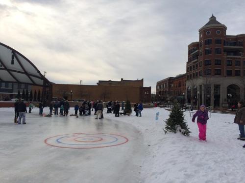 A winter scene with people skating on an ice rink, surrounded by snow and buildings, under a cloudy sky.