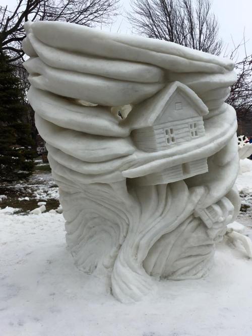 A whimsical snow sculpture featuring swirling snow with a small house embedded in it, set in a snowy landscape.
