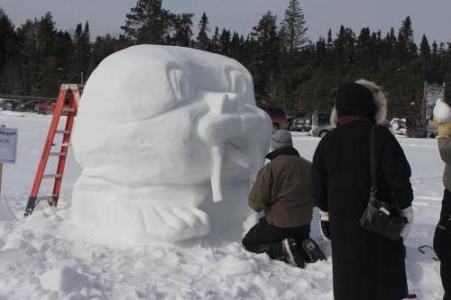 A large snow sculpture resembling a walrus, with people working on it in a snowy landscape. Trees in the background.