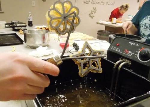 A hand holds a flower-shaped mold over a fryer, with golden pastries cooking in hot oil.