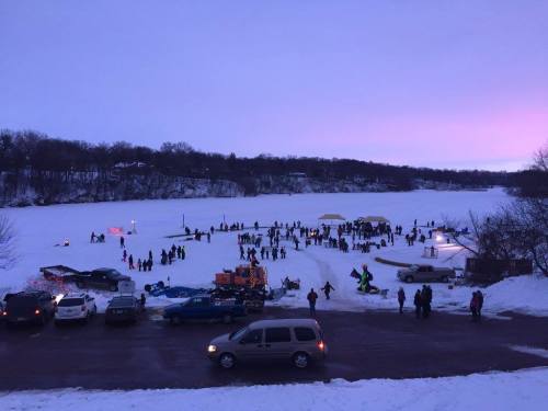 A snowy landscape with people gathered on a frozen lake, enjoying winter activities as the sun sets in the background.