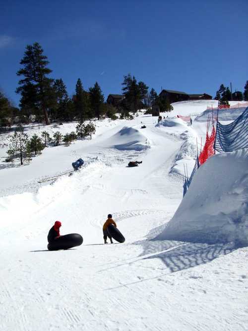 Two people in winter gear are tubing down a snowy slope, surrounded by trees and a clear blue sky.