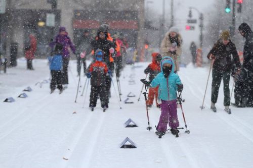 Children and adults ski on a snowy street, with snowflakes falling and colorful winter clothing visible.