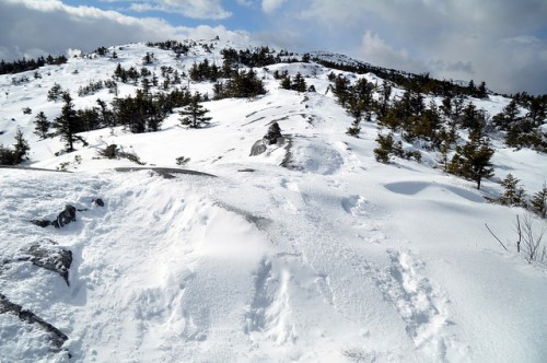 A snowy landscape with rocky terrain and scattered trees under a partly cloudy sky.
