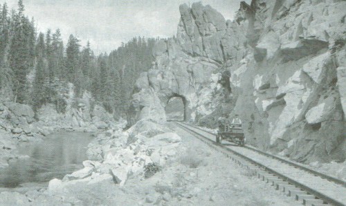 Black and white image of a rocky landscape with a train track and a vintage car near a river and forested hills.
