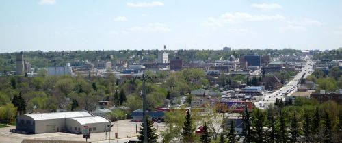 A panoramic view of a city surrounded by greenery, featuring buildings and a busy road under a clear blue sky.