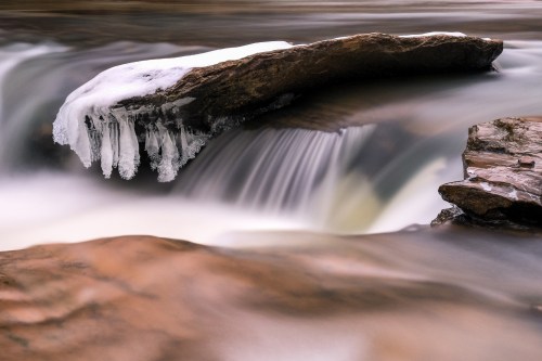 A serene river scene featuring a rock with icicles, flowing water, and smooth stones in a tranquil setting.
