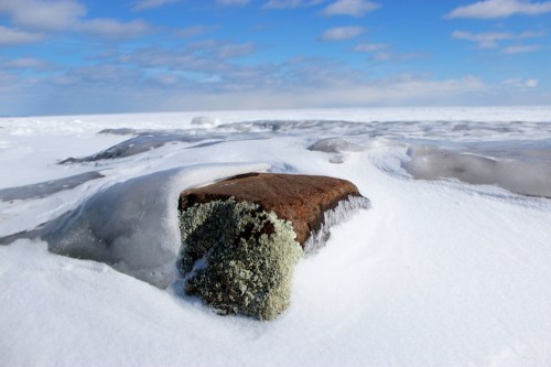 A rocky outcrop covered in snow and ice, with a clear blue sky and scattered clouds in the background.